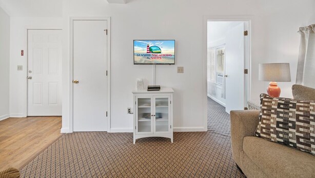 A living room with a television on the wall, a white cabinet, a patterned carpet, a sofa, and a door leading to another room. - Dolly's Place 6 by Seaside Vacations! (Chincoteague Island)