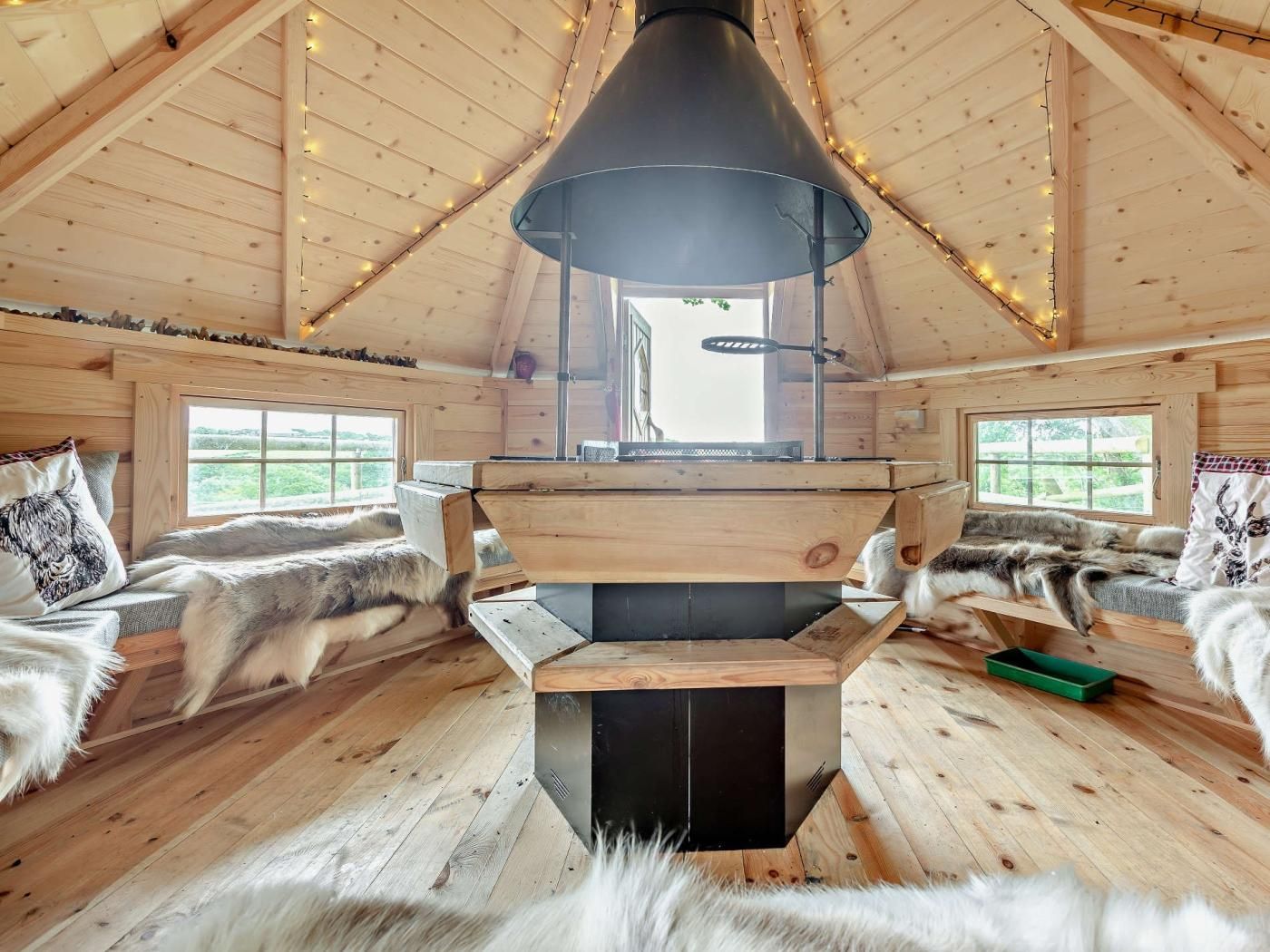 A cabin interior with a central wood-burning stove, wooden walls, and fur-covered furniture.
