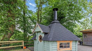 Eine kleine, hellgrüne Holzhütte mit einem schwarzen Schornstein, einer Holzterrasse und einem eingezäunten Bereich.