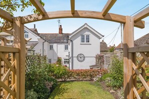 A white house with a clock on the wall, surrounded by a garden with various plants and a wooden gate. - 3 Bed in Bridport (oc-dc055) (Burton Bradstock)