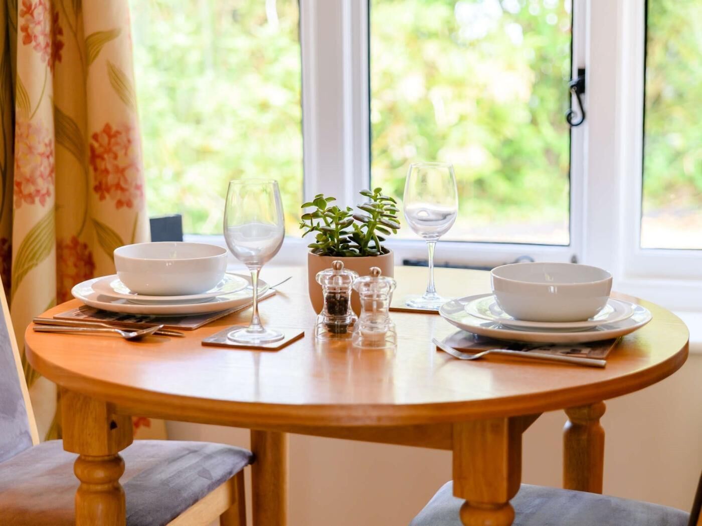 Une table ronde en bois, dressée pour le dîner, avec deux bols, deux verres à vin, une petite plante en pot et des salières et poivrières.