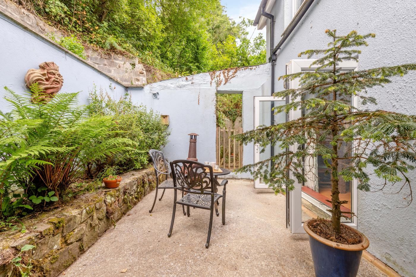 A courtyard with a table and chairs, surrounded by greenery and a stone wall.