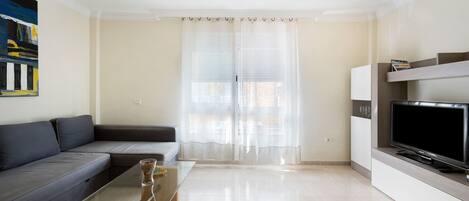 A modern living room with a sofa, a glass coffee table, a flat-screen TV, and sheer curtains.