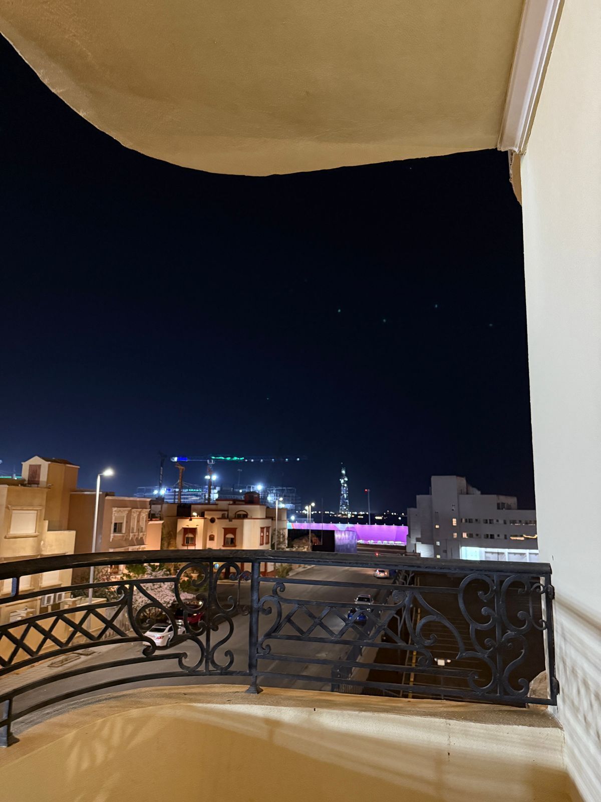 Een balkon met een metalen hekwerk, uitzicht op de verlichte skyline 's nachts, en gebouwen met gloeiende ramen.
