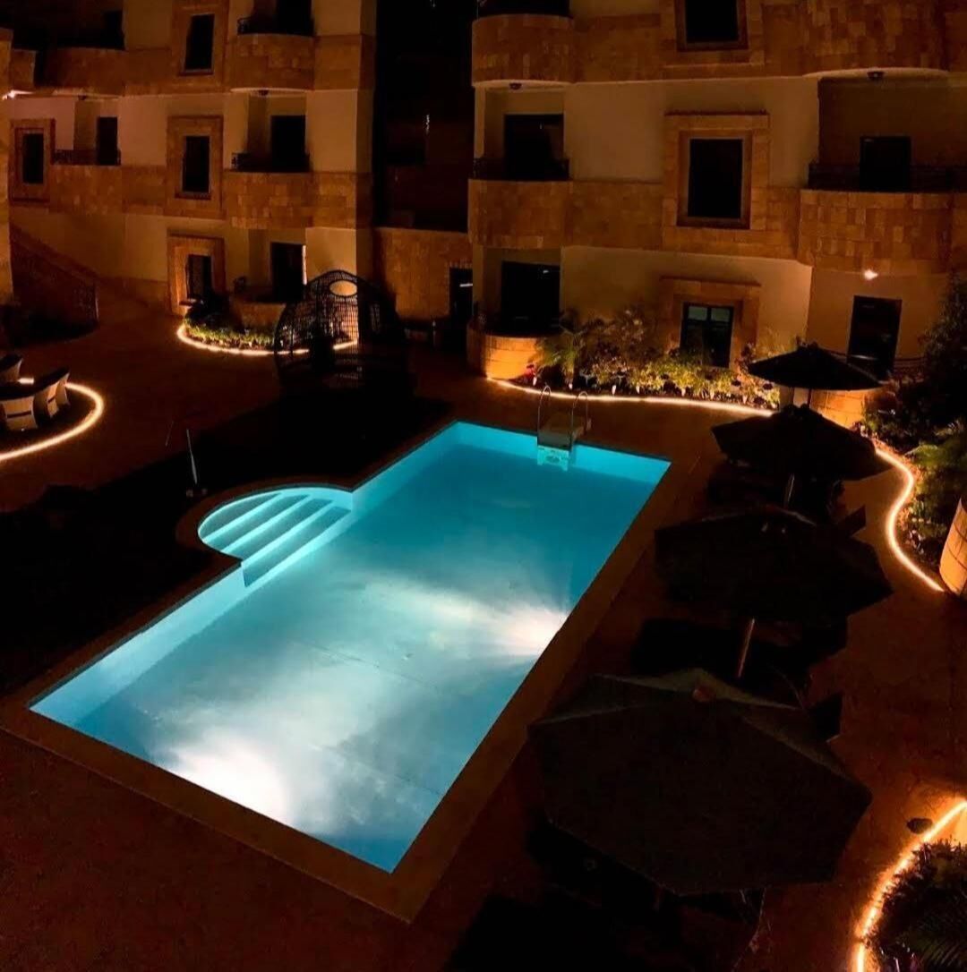 A swimming pool with a sunken seating area, surrounded by outdoor furniture and a building with multiple balconies.