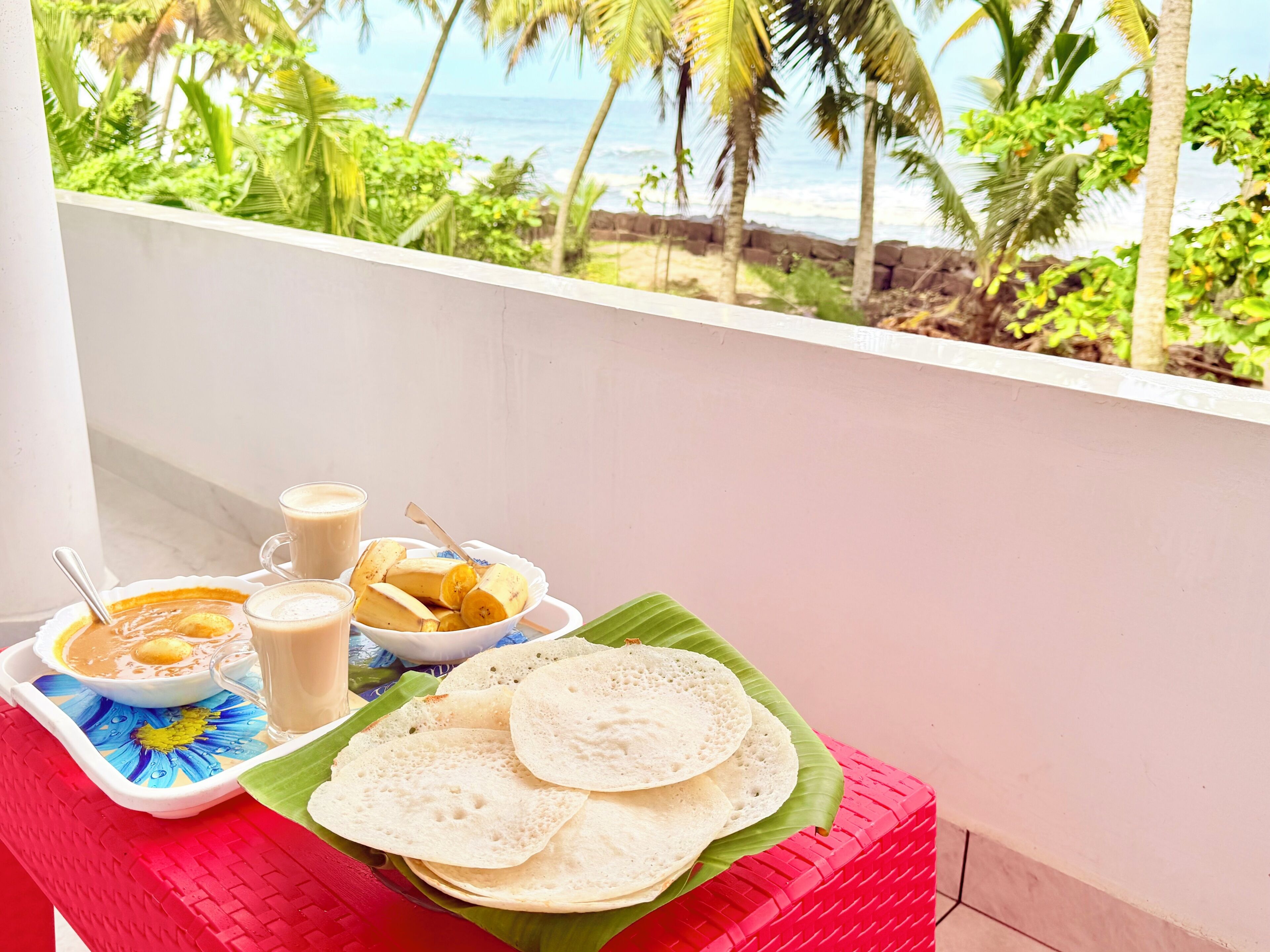 A balcony with a table set for a meal, including a bowl of soup, a plate of banana slices, and two glasses of a beverage.