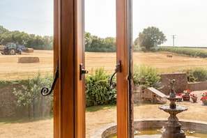 Vista a través de una ventana con marco de madera, una pared de piedra, una fuente y un campo con fardos de heno y un tractor.