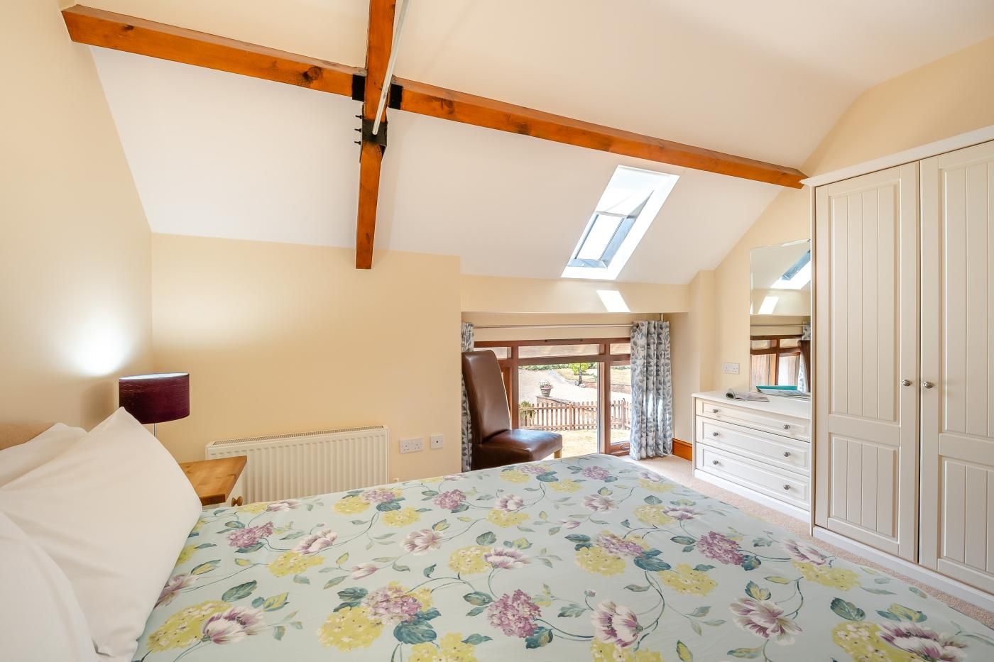 A bedroom with a sloped ceiling, a skylight, a bed with floral bedding, a dresser, and a chair.