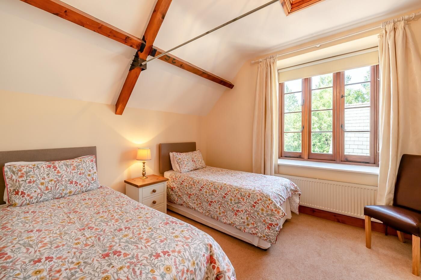A bedroom with two beds, a nightstand, a lamp, a window with curtains, and a radiator.