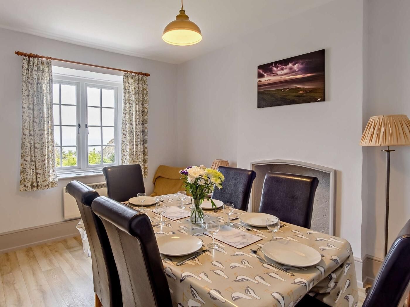 A dining room with a table set for four, a vase of flowers, and a painting on the wall.