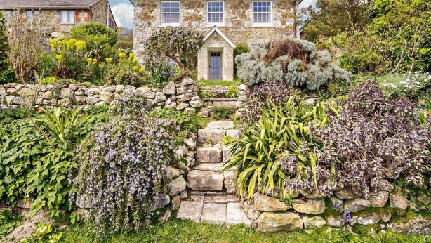 A stone house with a central door, surrounded by a garden with various plants and a stone wall. - 3 Bed in Niton Undercliff (oc-ic125) (Niton)