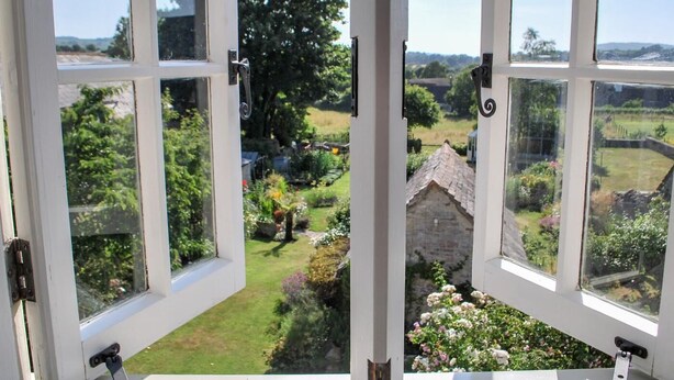Blick durch ein Fenster mit weißen Rahmen, das einen Garten mit einem Steinhaus, Bäumen und blauem Himmel zeigt.