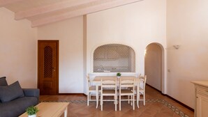 Un salon avec un canapé, une table basse ornée d’une plante, une table à manger avec des chaises, une porte en bois et un sol carrelé.