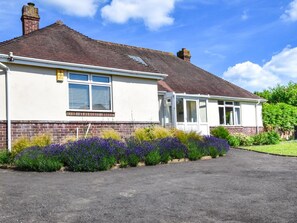 A single-story house with a brick facade, a tiled roof, and a front garden with purple flowers. - 3 Bed in Totland Bay (oc-ic110) (Totland)