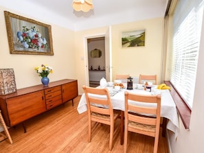 A dining area with a table set for two, a wooden cabinet, a vase with flowers, and a painting on the wall. - 3 Bed in Totland Bay (oc-ic110) (Totland)