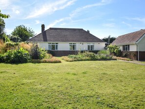 A single-story house with a sloped roof, a brick foundation, and a front yard with a lawn and various plants. - 3 Bed in Totland Bay (oc-ic110) (Totland)