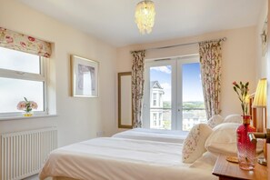 A hotel room with a bed, a bedside table with a lamp, a window with curtains, a radiator, and a view of a building outside. - 3 Bed in Shanklin (oc-ic113) (Shanklin)