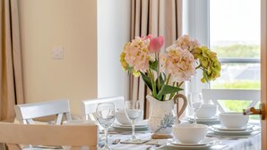 Un tavolo da pranzo apparecchiato con piatti bianchi, ciotole e bicchieri da vino, con un vaso di fiori al centro.