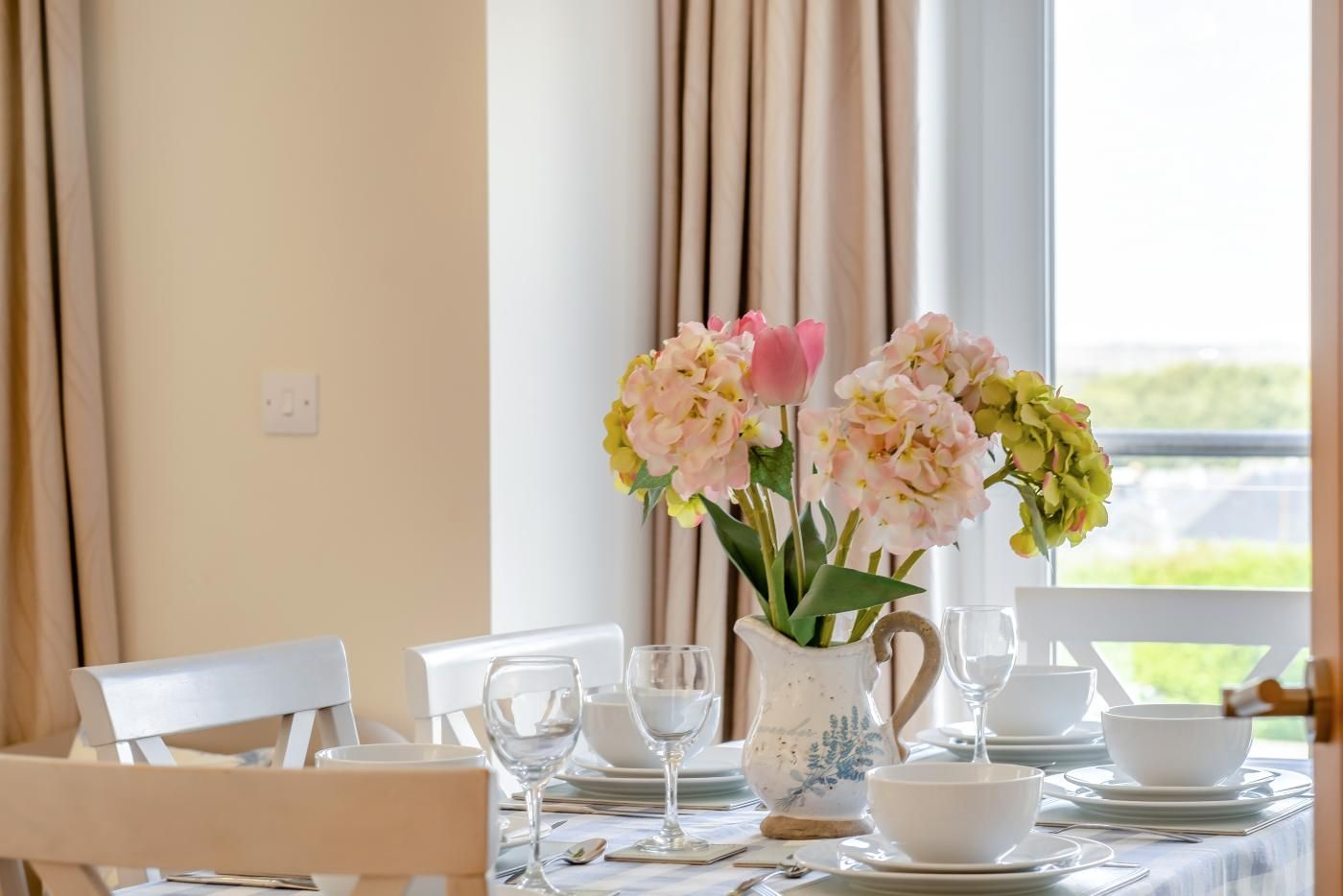 Une table à manger dressée avec de l’assiette blanche, des bols et des verres à vin, et un vase de fleurs placé au centre.
