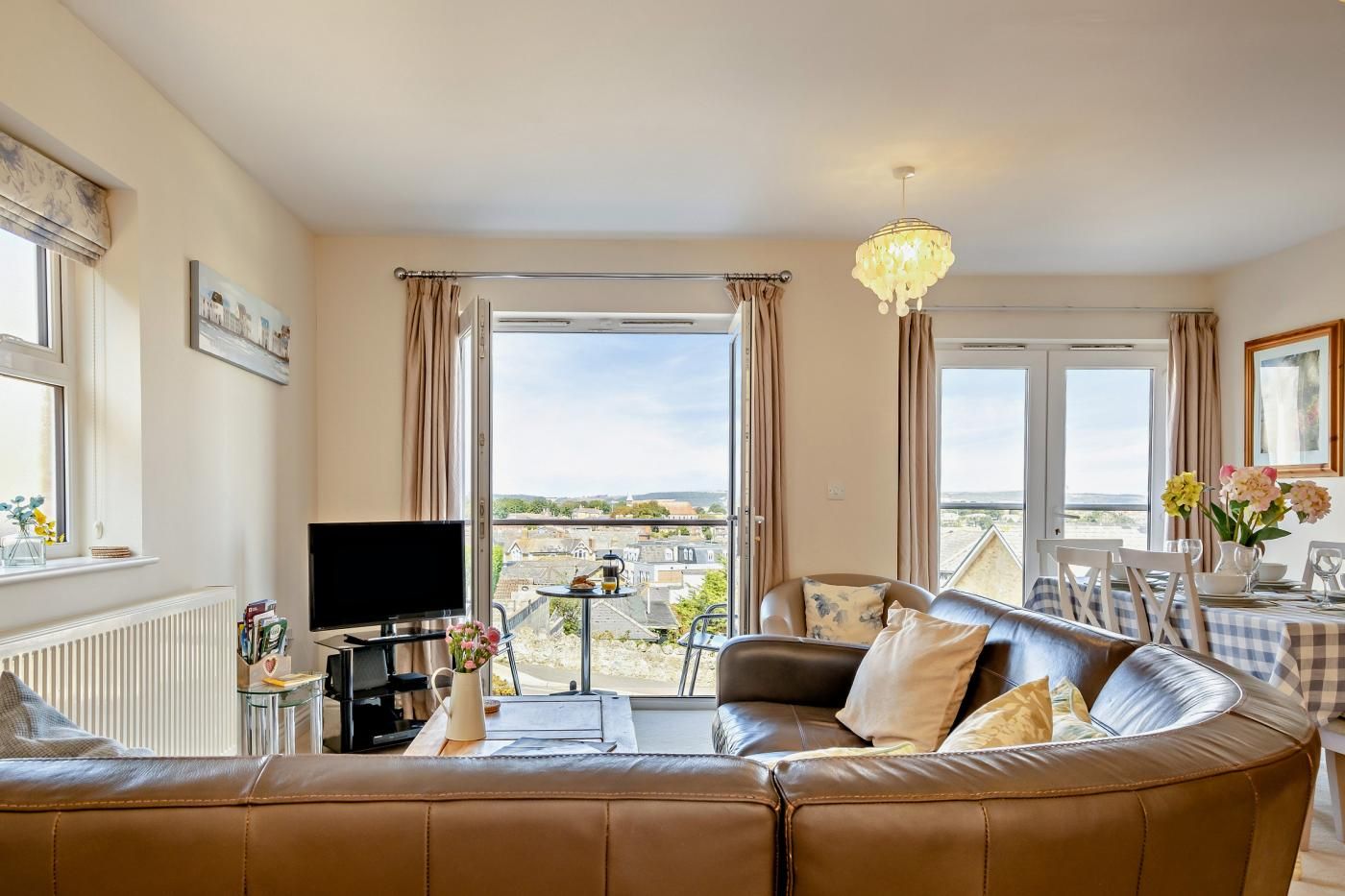 A living room with a leather sofa, a television, a dining table with chairs, and a view of the city through large windows.