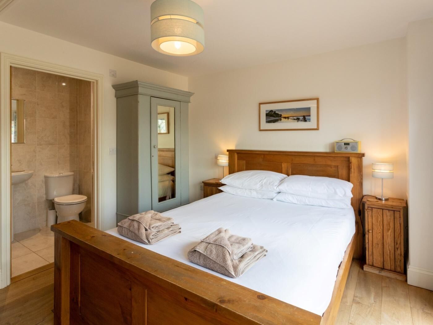 A bedroom with a wooden bed, a nightstand with a lamp, a bedside table, a wardrobe, and a bathroom with a sink and toilet.