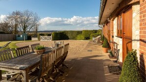 A patio with a wooden table and chairs, a brick building, and a view of the countryside. - 4 Bed in Bembridge (oc-ic130) (Hillway)