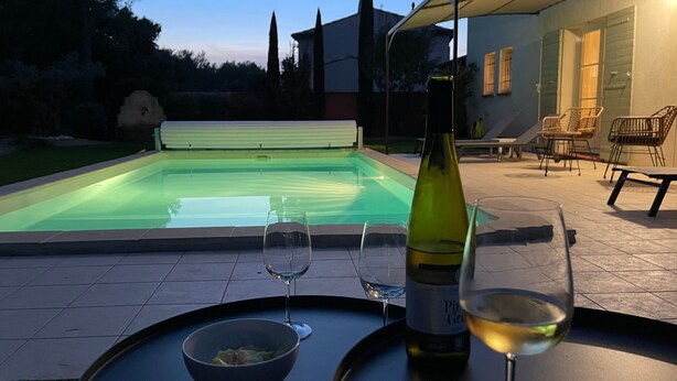 Une piscine avec un espace détente, une bouteille de vin et deux verres sur une table.