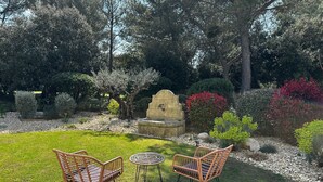 Un jardin agrémenté de deux fauteuils en osier, d’une fontaine en pierre et de divers arbustes.