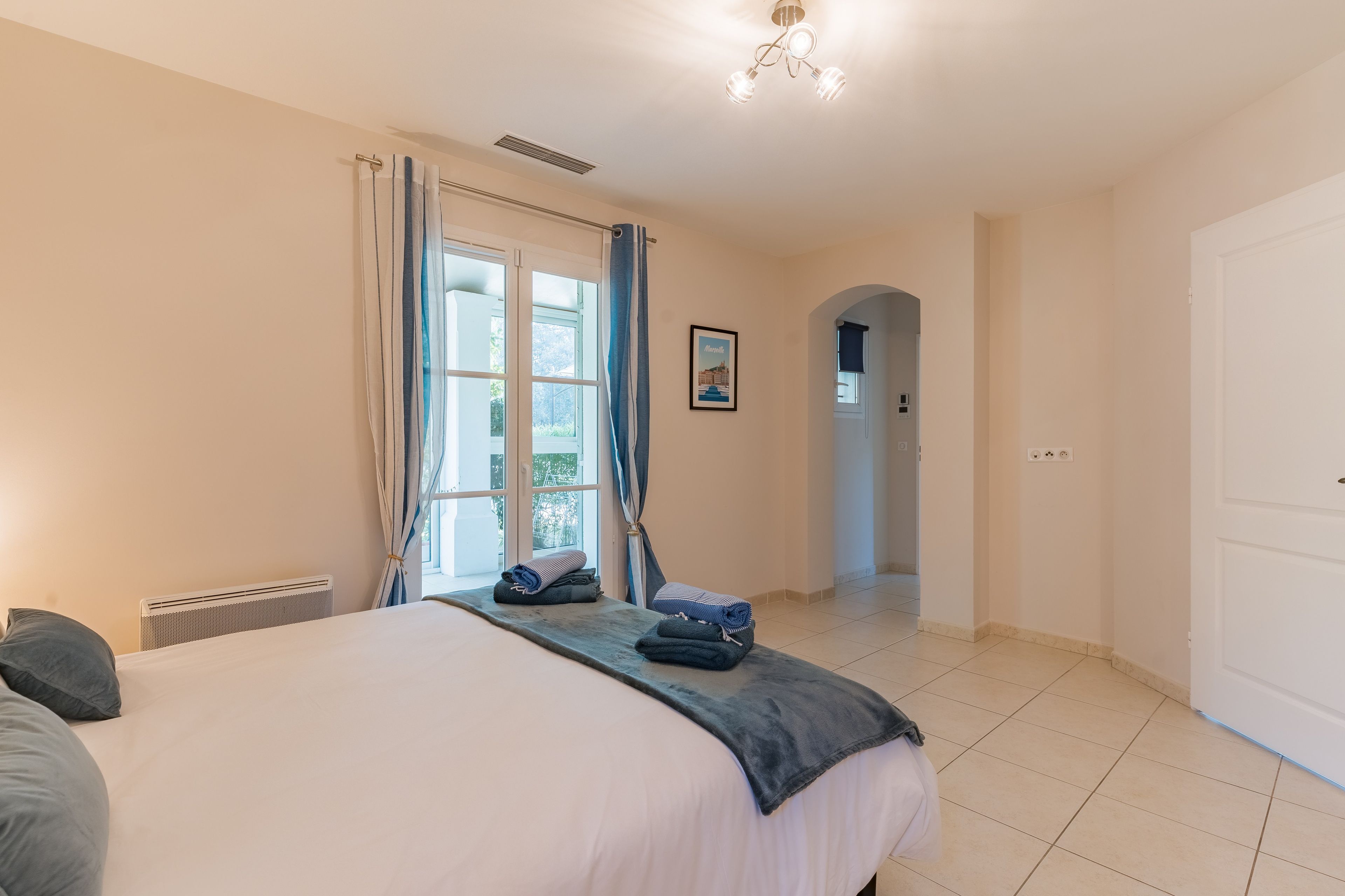 Une chambre d’hôtel avec un lit, un radiateur, des rideaux, un tableau encadré et un couloir menant à une autre pièce.