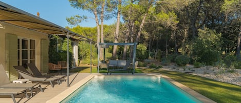 Una piscina con sillones de descanso y un pérgola, rodeada de árboles y un césped cuidado.