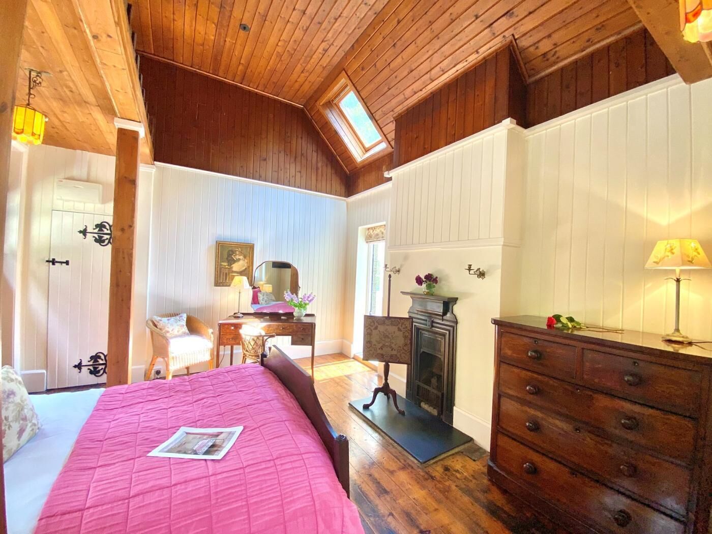 Ein Schlafzimmer mit schrägen Wänden, einem Bett mit rosa Decke, einem Schminktisch, einem Spiegel, einem Kamin und einem Dachfenster.
