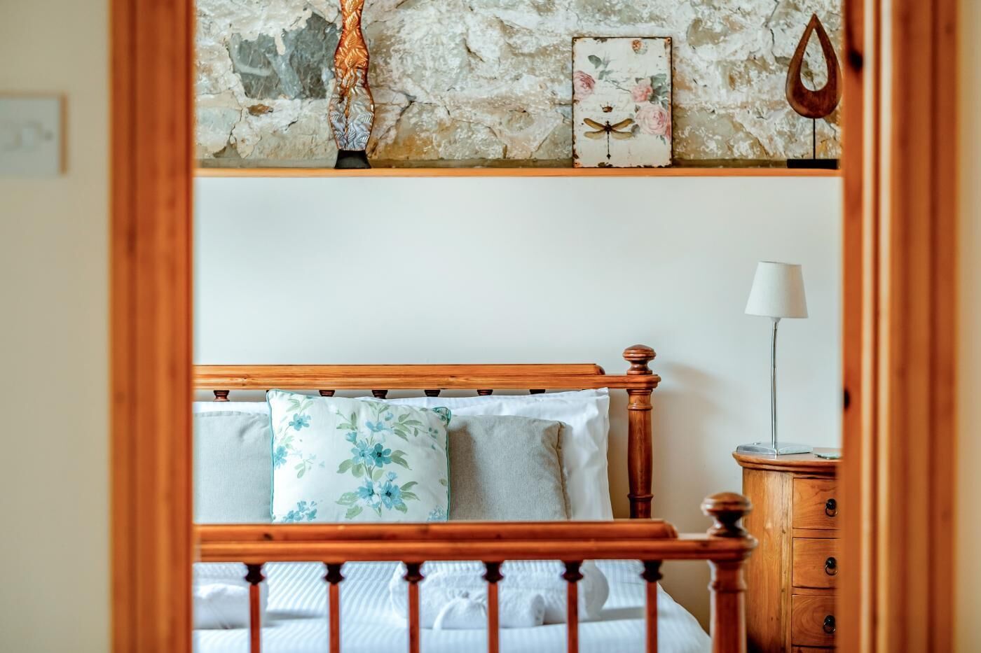 A bedroom with a wooden bed, floral pillows, a bedside lamp, and a rustic wall.