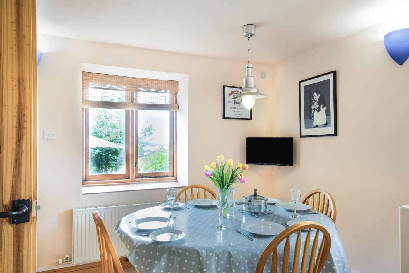 A dining area with a table set for four, a window with a view of greenery, a television mounted on the wall, and a framed picture.