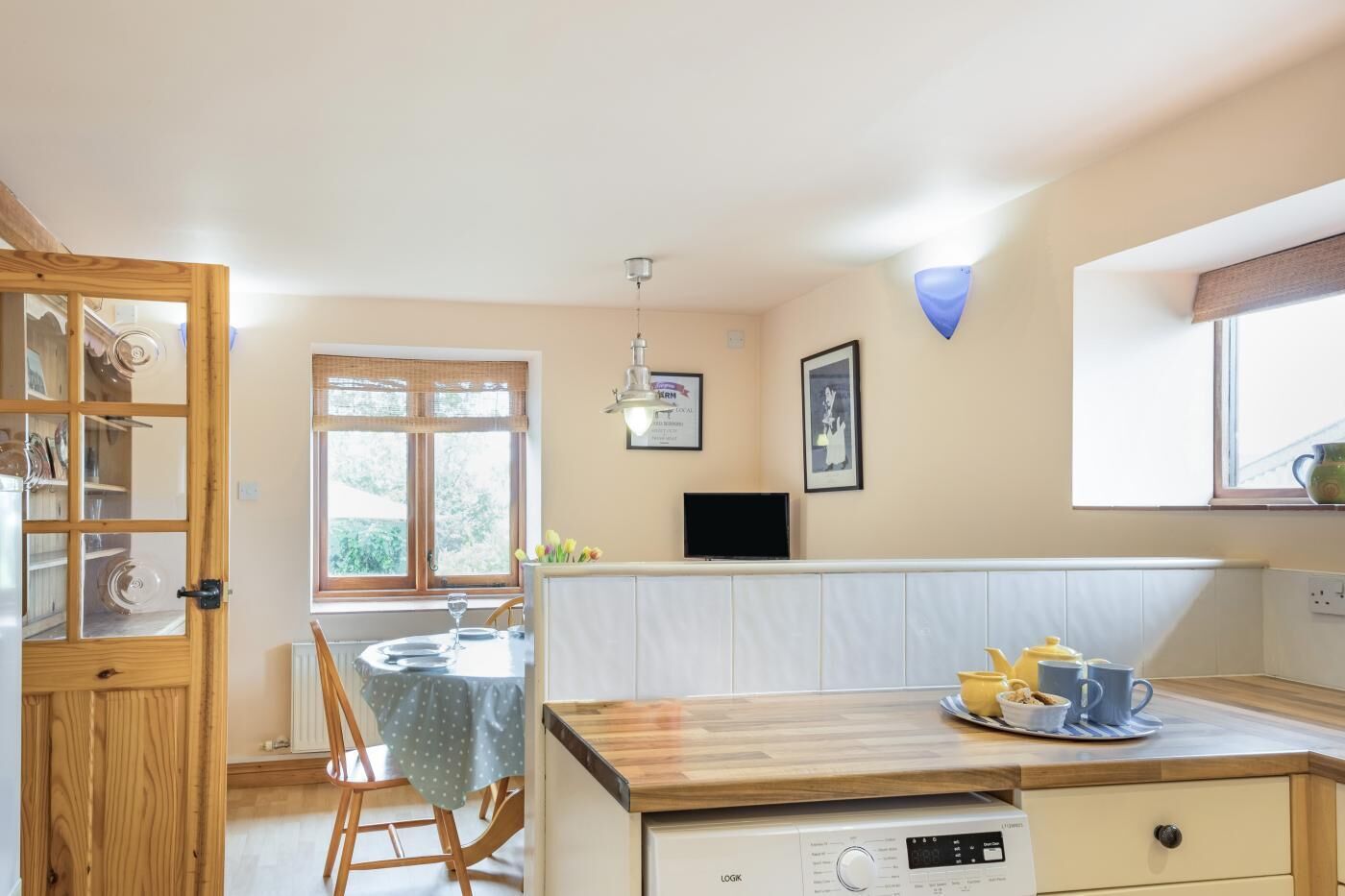 A kitchen with a wooden table, chairs, a dishwasher, a window, and a wall-mounted television.