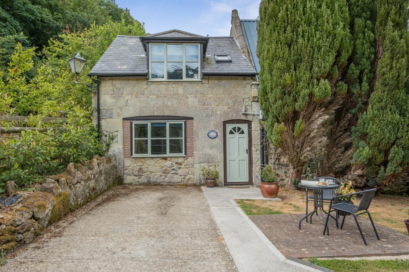A stone house with a gravel driveway, a front door, and a small patio with a table and chairs.