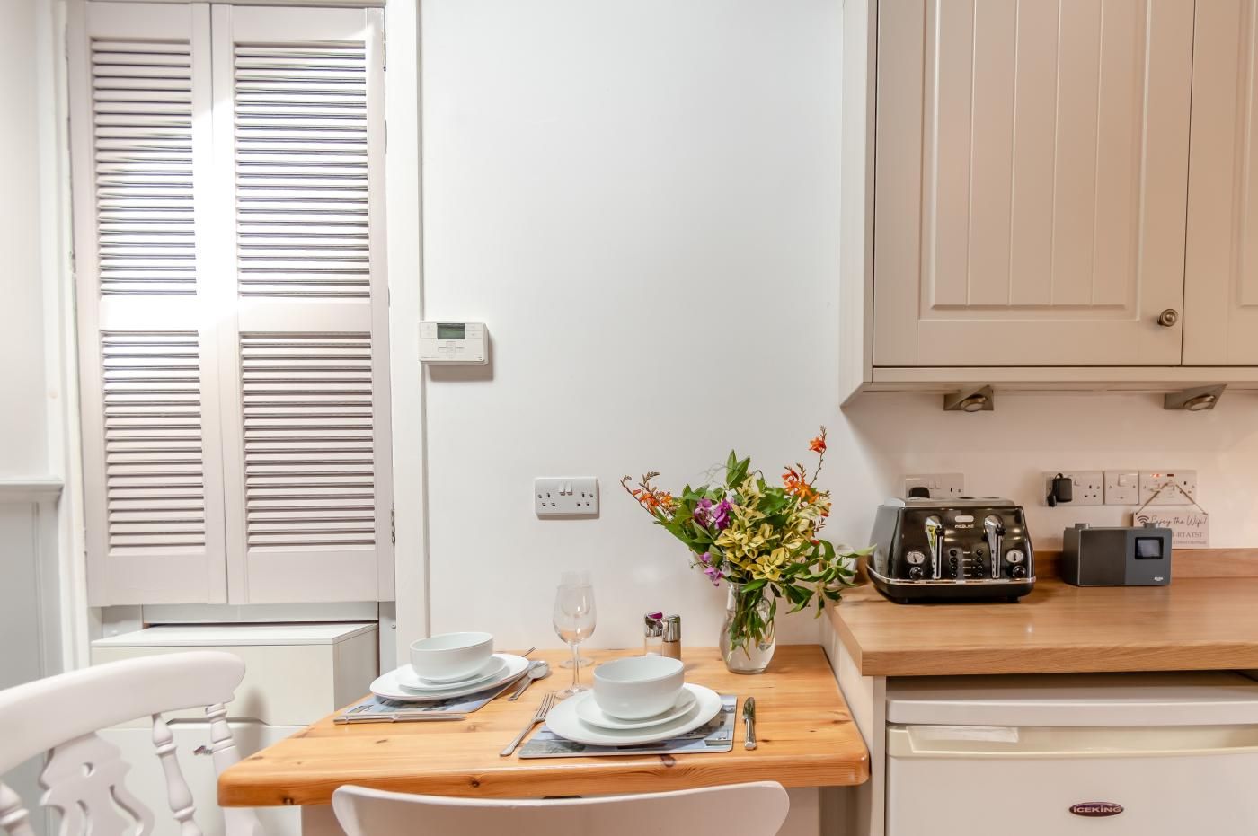 A dining area with a wooden table set for two, white chairs, a vase with flowers, a radio, and a kitchen area with cabinets and appliances.