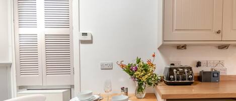 A dining area with a wooden table set for two, white chairs, a vase with flowers, a radio, and a kitchen area with cabinets and appliances.