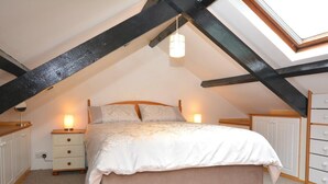 A bedroom with a sloped ceiling, a bed with a white duvet, two bedside lamps, a small dresser, and a colorful striped rug. - 1 Bed in Haverfordwest (oc-61170) (Porthgain)