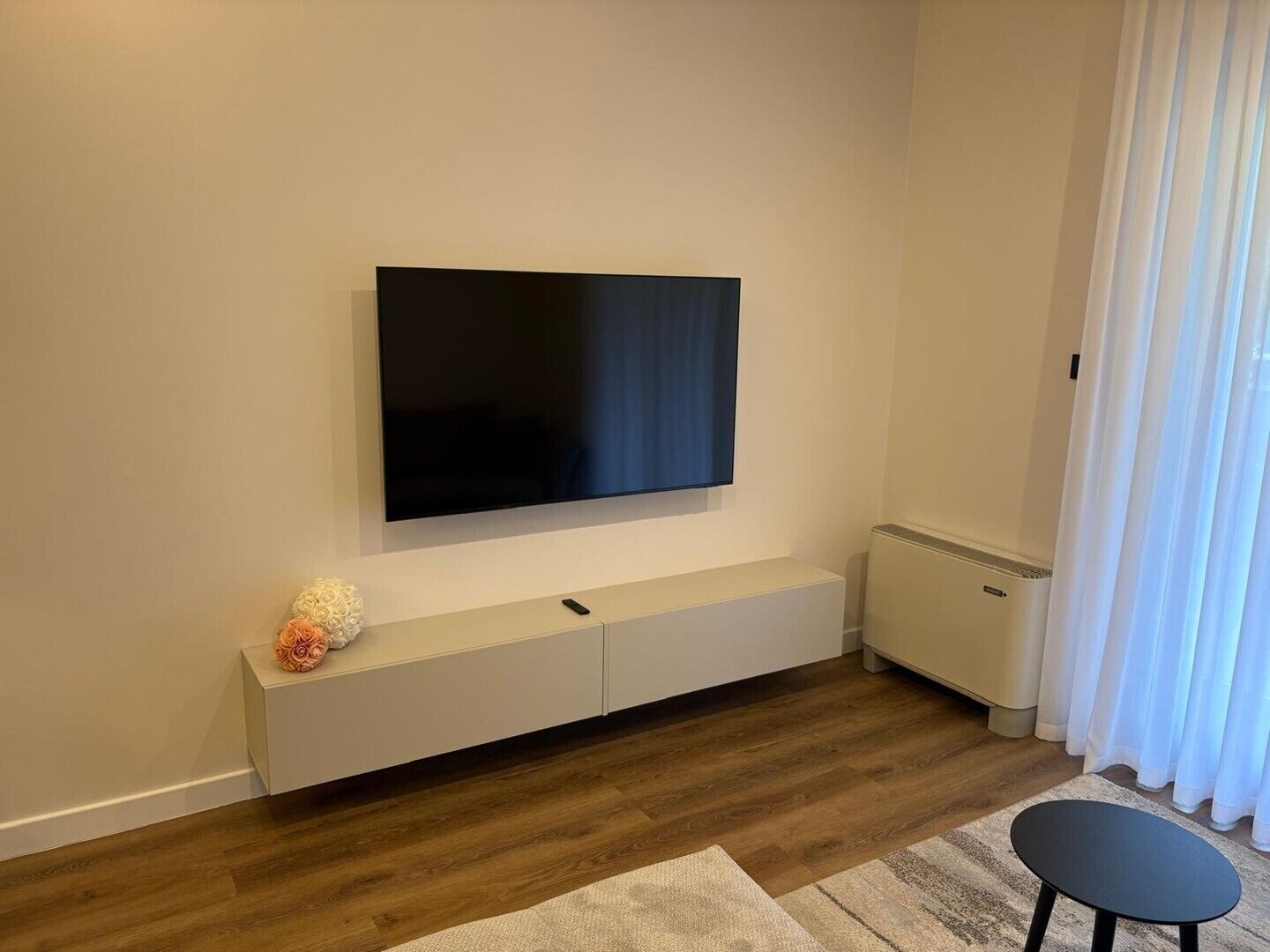 Ett modernt vardagsrum med en platt-TV som är monterad på väggen, en vit TV-bänk, ett litet bord och ett fönster med vita gardiner.