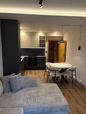 A modern living room with a sofa, a dining table with chairs, a kitchen area with cabinets, and a pendant light. - Apartment Villa Milčetić (Malinska)