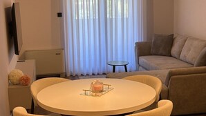 Olohuoneessa on pyöreä pöytä, sohva, televisio ja ikkuna, jossa on verhot.