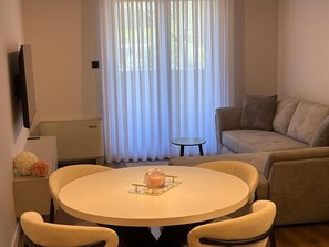 Ein Wohnzimmer mit rundem Tisch, einer Couch, einem Fernseher und einem Fenster mit Vorhängen.
