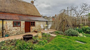 Una cabaña de piedra con techo de paja, un jardín con diversas plantas y una silla blanca al aire libre.