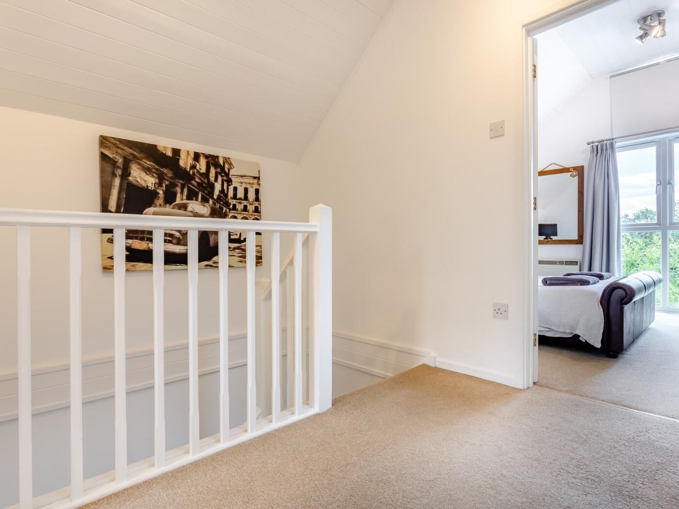 A hallway with a white railing, a painting on the wall, and a glimpse of a bedroom with a bed and curtains.