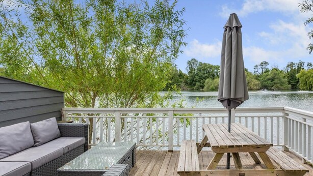 A deck with a sofa, a table, and an umbrella, overlooking a lake with trees.