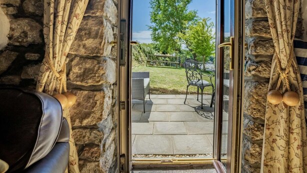 Un muro in pietra con una porta vetrata che dà su un patio esterno con prato, un albero e mobili da giardino.