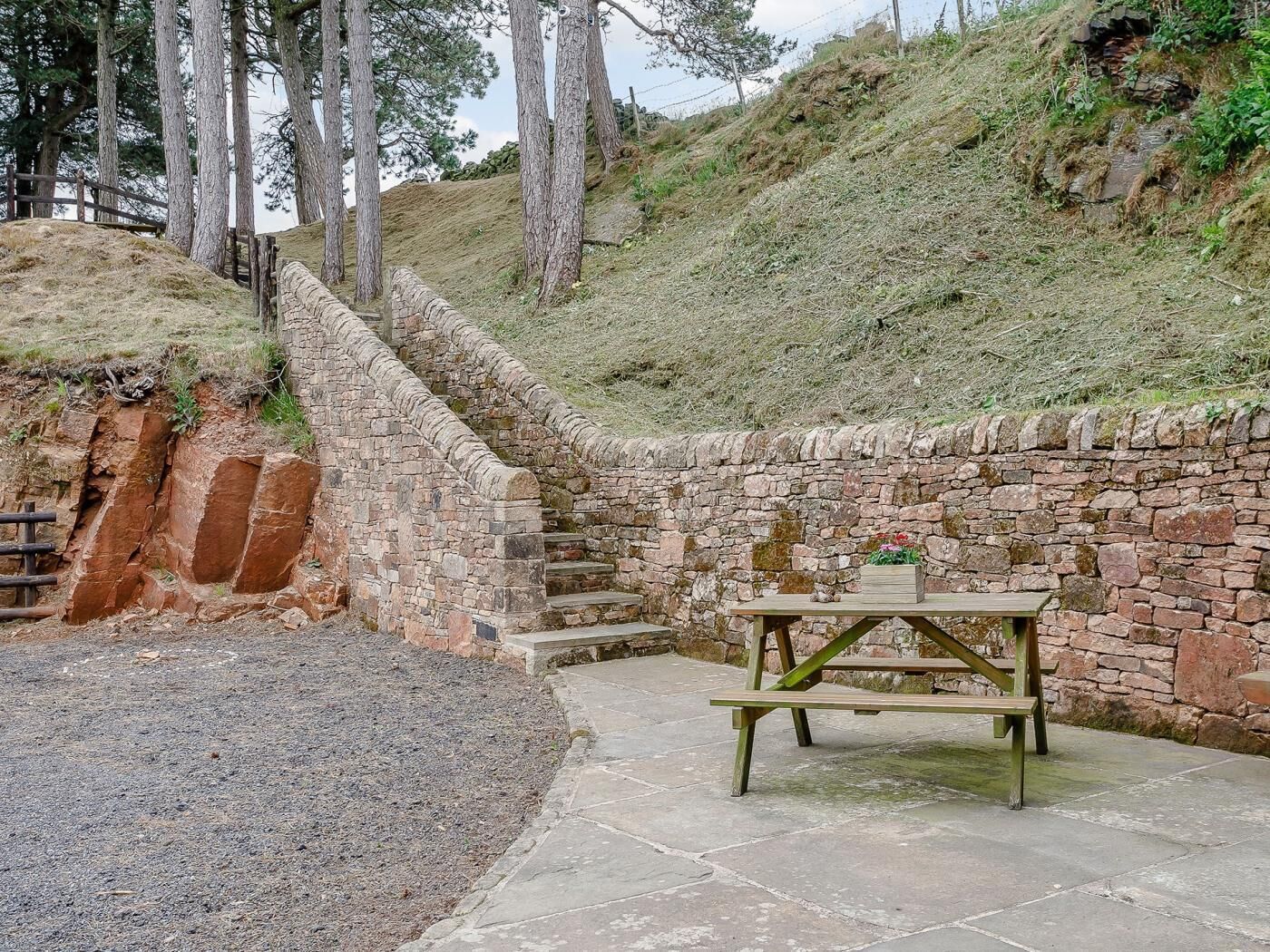 Mur de pedra amb taula de pícnic i bancs, envoltat d'una zona de grava i arbres.