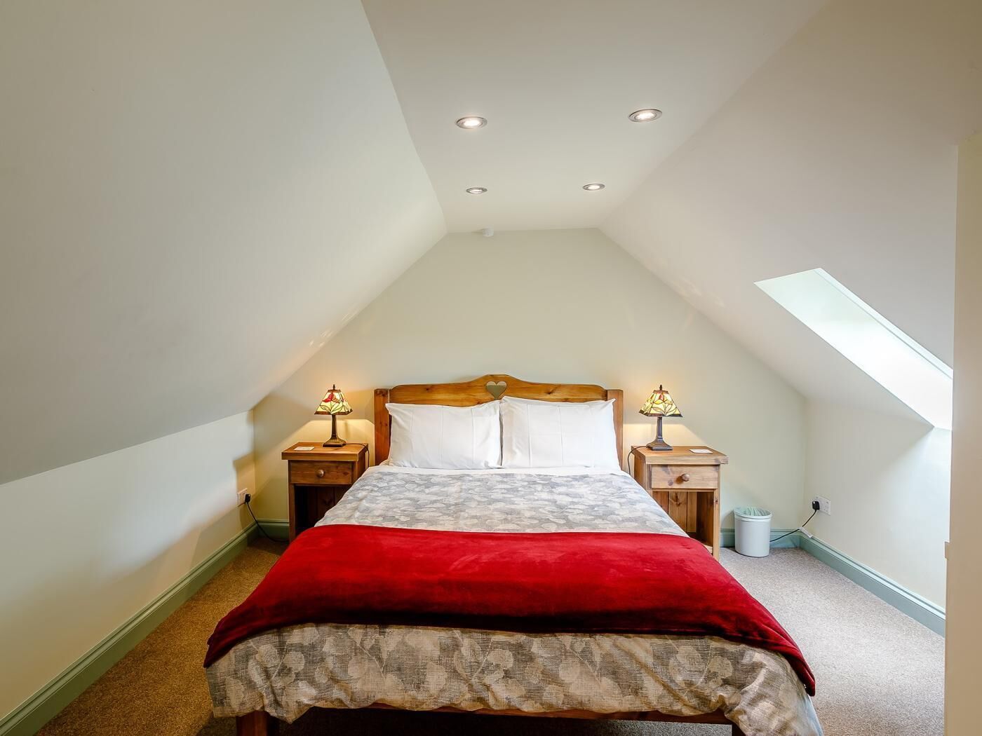 Een slaapkamer met een schuine plafond, een bed met een rode deken, twee bedlampen en een dakraam.