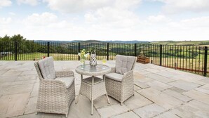 A patio with wicker furniture, a round table, and a view of rolling hills. - 4 Bed in Denbigh (oc-62368) (Denbigh)