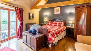 Une chambre avec un plafond en pente, un lit recouvert d’une couverture à carreaux, une table de chevet en bois, une commode et une fenêtre donnant sur une terrasse.